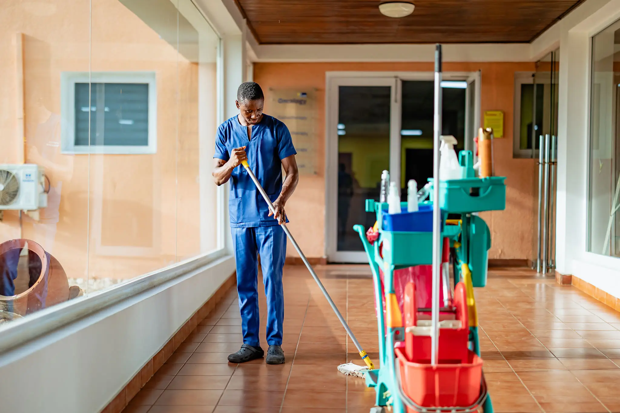 A janitor mopping a hospital corridor with cleaning equipment nearby.